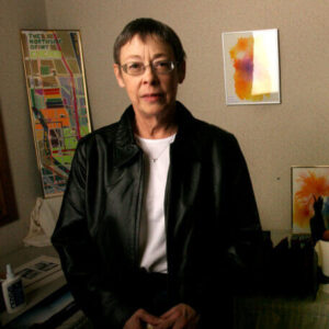 Julia Kellman in a black leather jacket stands indoors with colorful art and a map on the wall behind her.