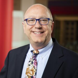 Sean Ulmer in a suit, glasses, and floral tie, standing indoors with a blurred background.